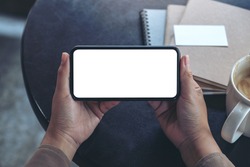 Top view mockup image of hands holding and using a black mobile phone with blank screen horizontally for watching with coffee cup and notebooks on table