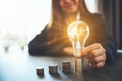 Businesswoman holding and putting lightbulb on coins stack on table for saving energy and money concept