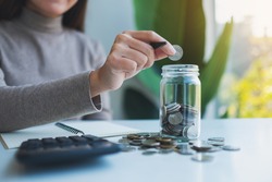 Closeup image of a woman putting coins in a glass jar with calculator on the table for saving money and financial concept