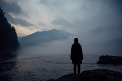 Alone human silhouette in white haze against the mountains and river. Thick fog in the evening twilight. Mysterious atmosphere. Reflection, meditation.