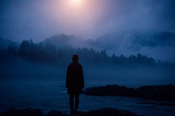 Dark human silhouette in a thick fog against the background of forest, hills and mountain river. Mysterious female figure on desert shore. Apocalyptic landscape, atmospheric pollution.