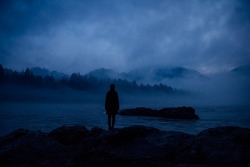 Human silhouette in a thick blue fog on the background of wooded hills and mountain river. Lonely female figure on a mysterious shore. Contemplation, meditation, unity in nature.