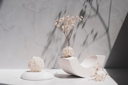 White geometric shape and podium, decorative balls and gypsophila flowers on a gray background. Modern interior still life with sunlight and shadows. 