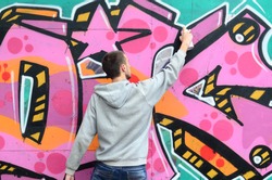 A young guy in a gray hoodie paints graffiti in pink and green colors on a wall in rainy weather