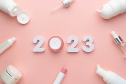 Best skincare products and cosmetic trends of 2023 concept. 2023 white number with lip balm, cream bottle, serum and lotion on pink background.