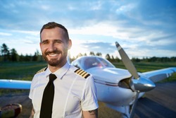 Cool young pilot with tattoos posing at small private motor airplane standing on runway at bright sunset. Blue sky, bright sun