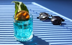 Drinks and cocktails. Blue curacao in a transparent glass with an orange chip with ice and a leaf on a bright blue background. Sunglasses, sunny day, summer. Background image, copy space. Minimalism