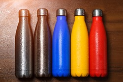 Colorful stainless thermo bottles, on a wooden table sprayed with water. Matte red bottle, blue, yellow and platinum color. With sunlight effect.