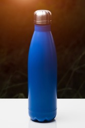 Stainless thermo bottle for water, tea and coffe, on white table. Dark grass background with sunlight effect. Thermos blue color.