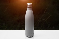 Stainless thermo bottle for water, tea and coffe, on white table. Dark grass background with sunlight effect. Thermos silver color.