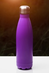 Stainless thermo bottle for water, tea and coffe, on white table. Dark grass background with sunlight effect. Thermos violet purple color.