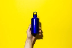 Male hand holding aluminium thermo eco water bottle, classic blue of color, isolated on yellow background.