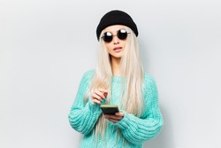 Studio portrait of young hipster blonde girl using smartphone on white background. Wearing round sunglasses and black beanie hat.