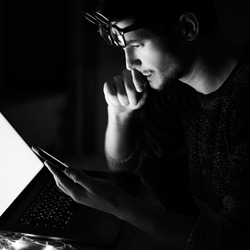 Black and white photo; portrait of thoughtful young man wearing glasses using smartphone and laptop.
