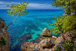 Beautiful turquoise and blue water near rocks and cliffs in Negril, Jamaica. A tree on the left and rocks on the right with a beautiful lagoon in the sea