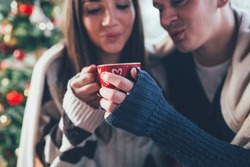 Young couple drinking hot tea together, with a Christmas tree in a background. Selective focus on man's hand.