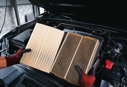Auto mechanic compares a clean new air filter to an old, filthy one with a engine compartment background
