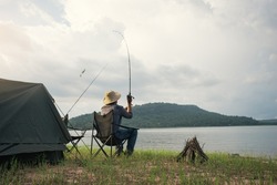 Angler sitting a chair and cast rod fishing at the lake. Fisherman with camping tent and bonfire for cooking on the shore of the lake. Survival concept.
