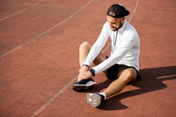 male runner in white black clothes holds his knee at the stadium, sports injury