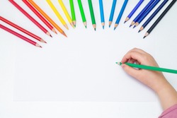 Top view of kid's left hand holding color pencil and drawing on white blank paper with the set of colour pencils. Copy space for text. Mockup.