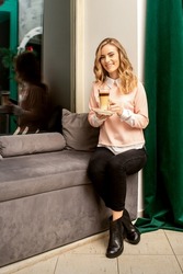 Beautiful caucasian young smiling woman with long wavy blonde hair holding and drinking a latte from a glass cup, sitting on a sofa