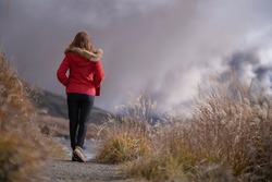 Asian woman in red sweater and long black legging walking, heading to the mountain with volcano smoke along the path with fountain grass. Traveller walking to volcano.