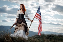 Girl in historical dress of 18th century with flag of United States. July 4 is US Independence Day. Woman of patriot freedom fighter in outdoor on background cloudy sky
