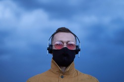 Cute serious caucasian male in pink eyeglasses, black protective medical face mask and orange jacket with headphones listening to the music, new album or soundtrack. Blue rainy sky weather