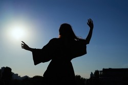 Dancing silhouette of attractive caucasian elegant girl in dress at sunset or woman performing modern dance outdoor in sunny day against blue sky. Freedom or success concept. High quality image