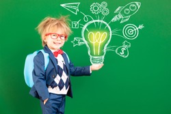 Bright idea! Funny child student with backpack in class. Happy kid against green chalkboard. Online education and e-learning concept. Back to school