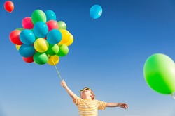 Happy child playing with bright multicolor balloons outdoor. Kid having fun in green spring field against blue sky background. Healthy and active lifestyle concept