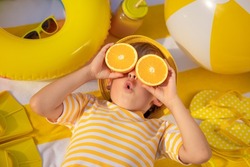 Surprized child holding slices of orange fruit like sunglasses. Kid wearing striped yellow t-shirt lying on beach towel. Healthy eating and summer vacation concept