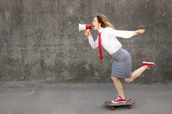 Happy businesswoman riding skateboard. Outdoor portrait of young woman shouting through loudspeaker. Back to work, start up and business idea concept
