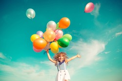 Happy child jumping with colorful toy balloons outdoors. Smiling kid having fun in green spring field against blue sky background. Freedom concept