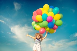 Happy child jumping with colorful toy balloons outdoors. Smiling kid having fun in green spring field against blue sky background. Freedom concept