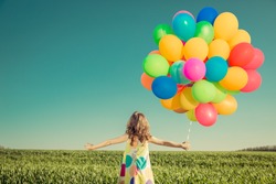 Happy child playing with colorful toy balloons outdoors. Little girl having fun in green spring field against blue sky background. Freedom and imagination concept