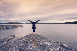 Freedom Happy Woman raised hands up to sky standing on the Island rock on the sea background with twilgiht colours in Sweden summer