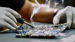 Technician engineer in workshop. Repairman in gloves is soldering circuit board of electronic device on the table, hands close up. He takes tin with a soldering iron and puts it on microcircuit.