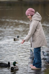 A little girl in a city park feeds the birds in a fashionable coat and hat. 