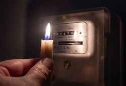 A man's hand with candle in complete darkness looking on electricity meter at home. Power outage, blackout concept. 