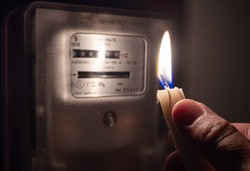 A man's hand with candle in complete darkness near an electricity meter at home. Power outage, blackout, non-payment of electricity bill concept.