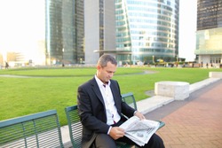 Happy male manager reading newspaper and smiling outside in  . Concept of businessman and mass media. Man dressed in black suit having break.