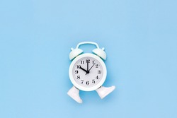 White alarm clock in toy sneakers walks on blue background. Minimal time movement concept. Place for text
