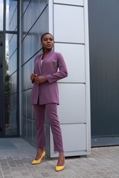 Outdoor full body length portrait of African girl with braids and make up wearing purple stylish pants suit and posing at urban minimalist grey background. Diversity fashion lifestyle concept.