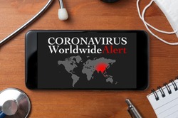 Coronavirus concept: a smartphone sorrounded by a stethoscope, a medical mask and other objects with a world map and an alert text on display