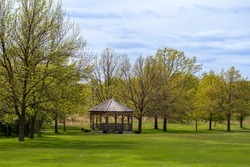 Landscape view of a  wooden gazebo in an ornamental botanical garden surrounded with green grass and trees, with blue sky