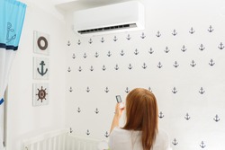 Portrait Of A Happy Woman Holding Remote Control In Front Of Air Conditioner At Home in childrens room.Air conditioner inside the cute baby boy’s room.With hot weather,is a good solution for family. 