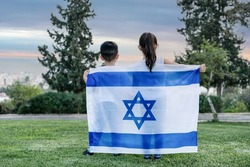 Little patriot Jewish children standing with the flag of Israel enjoying view of city. Back view image of young kids covering themselves with flag of Israel on meadow at sunset.