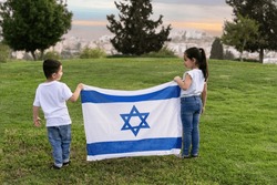 Young children stand on a hill holding an Israeli flag. A rear view little girl and boy with an Israel flag in the sunset.