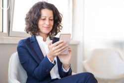 Satisfied customer using online mobile app. Business woman sitting in armchair, using mobile phone, looking at screen and smiling. Digital technology concept